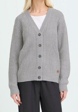 Femme portant un cardigan en maille gris clair boutonné sur une chemise blanche avec un pantalon noir, debout devant un fond uni.