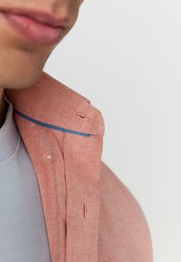 Nærbillede af en koralfarvet skjortekrave med en kontrasterende blå søm, der afslører et tekstureret stof og en knapdetalje nær halsen.