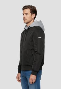 Giacca nera con cappuccio grigio, dotata di chiusura con zip, tasche laterali e polsini a costine. Il tessuto appare liscio e leggero.