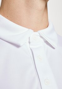 Hvid polo skjorte med klassisk krave, der har en glat tekstur og tre hvide knapper ved halsen. Nærbillede fokuserer på kravedesignet.