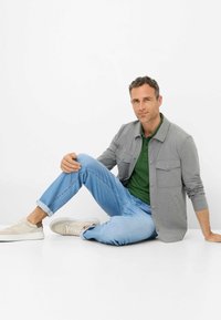 Giacca camicia grigia con chiusura a bottoni, polo verde, jeans blu e sneakers beige chiaro. Uomo seduto su uno sfondo bianco.