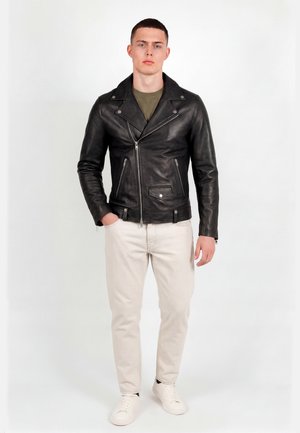 Young man standing wearing a black leather jacket, olive green shirt, light beige pants, and white sneakers, hands relaxed by sides.