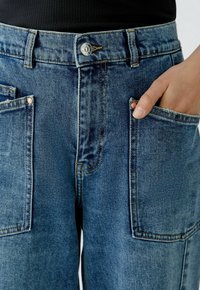 Jeans in mittlerem Blau mit hoher Taille, ausgestattet mit großen Fronttaschen, Kupferriegeln und kontrastierenden Nähten für zusätzliche Details.