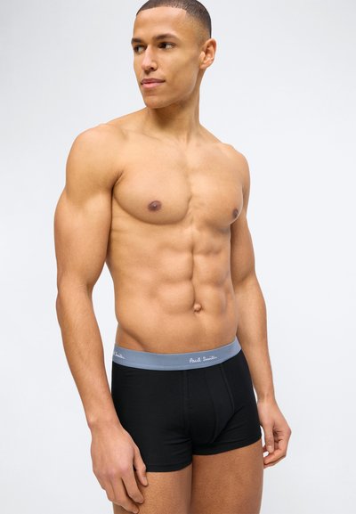 Μυώδης άνδρας που κοιτά προς τα αριστερά, φοράει μαύρα ανδρικά εσώρουχα με γκρι ζώνη μέσης με την επωνυμία "Paul Smith" σε λευκό απλό φόντο.