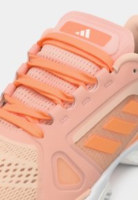 Κοντινή λήψη ενός αθλητικού παπουτσιού σε ροδακινί και κοραλλί χρώμα, με ύφασμα από δίχτυ, κορδόνια και τρεις πορτοκαλί ρίγες στο πλάι.