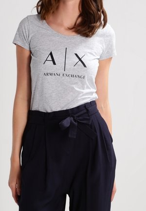 Nainen, jolla on vaaleanharmaa Armani Exchange -t-paita, joka on laitettu korkea vyötäröisiin mustiin housuihin ja sidottu kangasvyöllä.