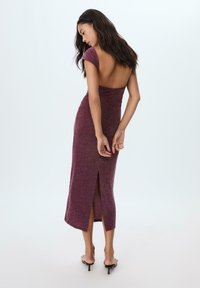 Donna che indossa un vestito midi prugna scintillante con schiena bassa e spacco, in piedi a piedi nudi su uno sfondo bianco, mani dietro la schiena.