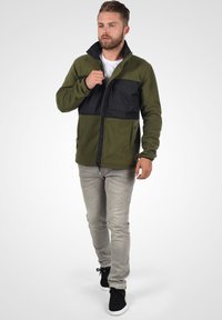 Fleece jacket in olive green with a black chest panel, full zipper, high collar, and two side pockets; worn over light jeans and black shoes.