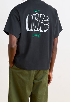 Personne portant un t-shirt noir Nike avec le texte blanc style graffiti "Nike" et le texte vert "Since '72" dans le dos, assorti à un pantalon vert.