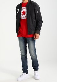 Schwarze Bomberjacke mit gerippten Bündchen, rotes T-Shirt mit grafischem Stern, blaue Jeans und weiße Turnschuhe.
