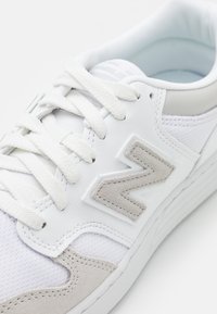 Bílé tenisky s bílými tkaničkami, šedými semišovými akcenty, síťovanou tkaninou a šedým stylizovaným logem "N" na boku.