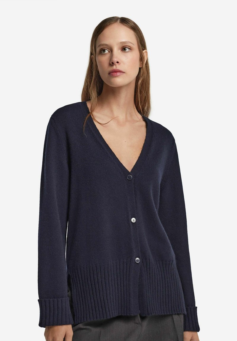 Kvinna i en marinblå V-ringad knappad cardigan med ribbade muddar och fåll, tillsammans med mörkgrå byxor, mot en vit bakgrund.