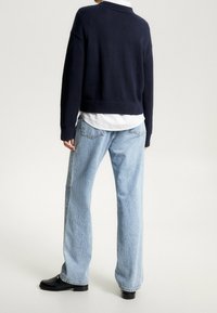 Eine Person steht mit dem Rücken zur Kamera und trägt hellblaue gerade Jeans, schwarze Loafers, ein weißes Hemd und einen dunkelblauen Pullover.