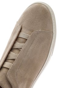 Beżowy zamszowy but z elastycznymi krzyżującymi się paskami i białą gumową podeszwą, pokazujący widoki z góry i z boku na przód i górną część.