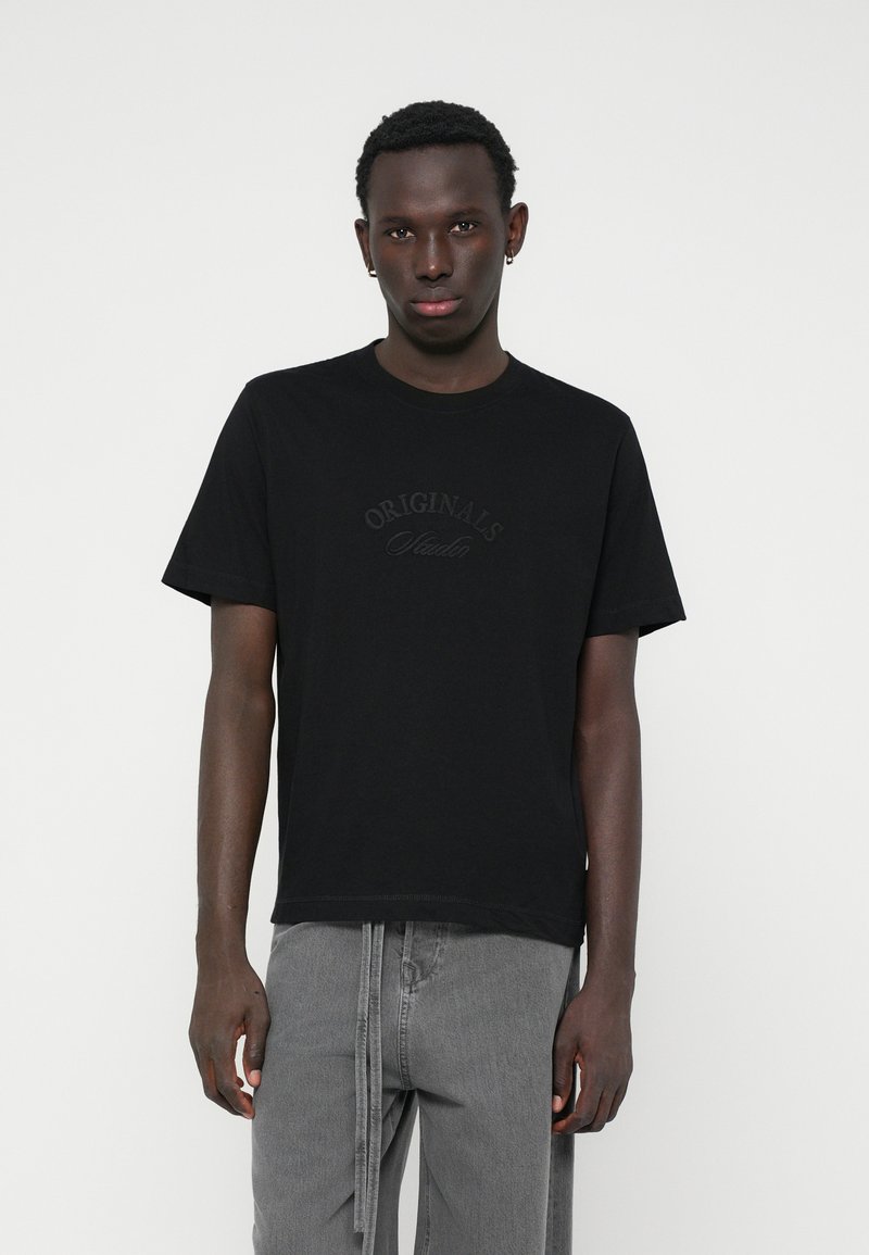 Camiseta de algodón negra de manga corta, con un sutil logo en relieve de "ORIGINALS" en el pecho. Combinada con pantalones grises.