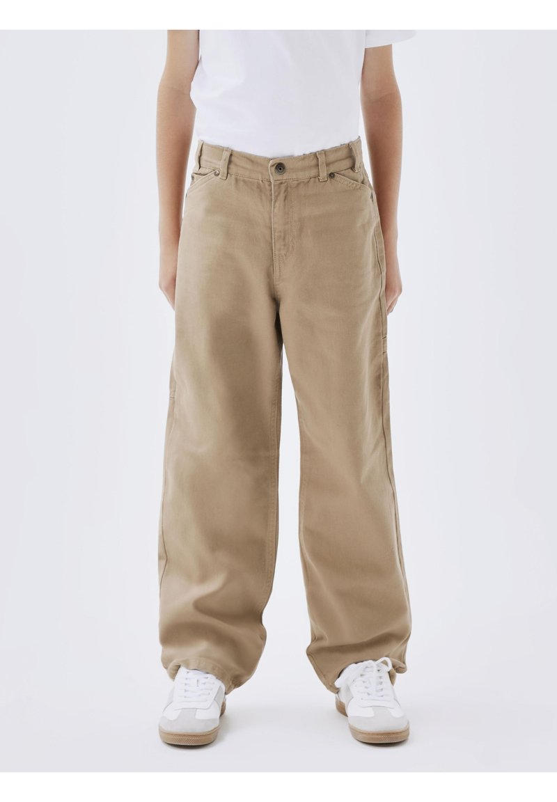 Personne portant un pantalon beige ample, des baskets blanches et une chemise blanche, debout devant un fond clair uni.