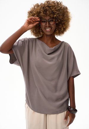 Uśmiechnięta osoba o kręconych włosach, poprawiająca duże okulary, ubrana w luźną szarą bluzkę, kremowe spodnie, złote kolczyki i ciemną bransoletkę.