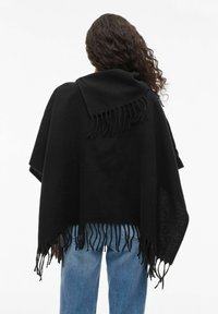 Person with long curly hair wears a black fringed shawl over blue jeans, viewed from the back against a plain light background.