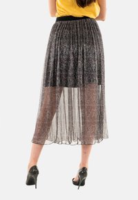 Jupe noire transparente avec un design plissé, un dos légèrement plus long et une taille élastique. Portée avec des chaussures à talons hauts noires.