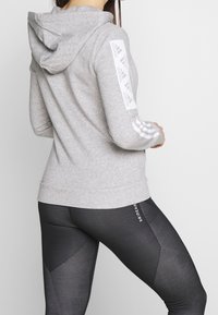 Felpa grigia con cappuccio e logo bianco Adidas, con strisce sulle maniche, abbinata a leggings neri testurizzati con motivi a contrasto.