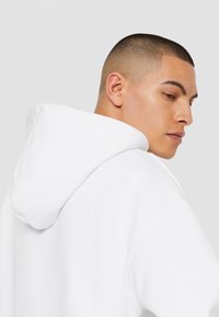 Hvid hætte sweatshirt med en blød tekstur, der har snoretræk i hætten og indsyede ærmer. Modellen vises fra en sidevinkel.