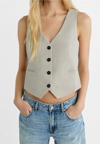 Grå ärmlös väst med djup V-ringning, fem svarta knappar, skräddarsydd passform och diskreta fickdetaljer, kombinerad med högmidjade jeans.