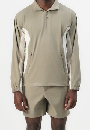 Mand iført beige langærmet trøje med lynlås og hvide sidelommer samt matchende beige shorts, stående foran en ensfarvet baggrund.