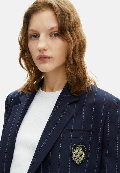 Blazer blu marino con righe sottili e distintivo ricamato in oro sulla tasca sinistra del petto, abbinato a un maglione bianco a coste indossato sotto.