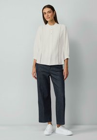 Chemise blanche à boutons avec un tissu texturé et un devant plissé, associée à un pantalon droit noir et à des baskets blanches.