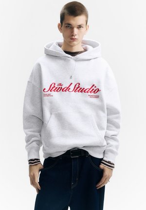 Ljusgrå hoodie med röd text, stor framficka, ribbade muddar och avslappnad passform, kombinerad med mörkblå jeans. Enkel design, avslappnad stil.
