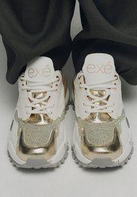 Zapatillas blancas con acentos de metal dorado y detalles iridiscentes, que cuentan con una suela acolchada y un diseño de cordones. Logotipo en la lengüeta.