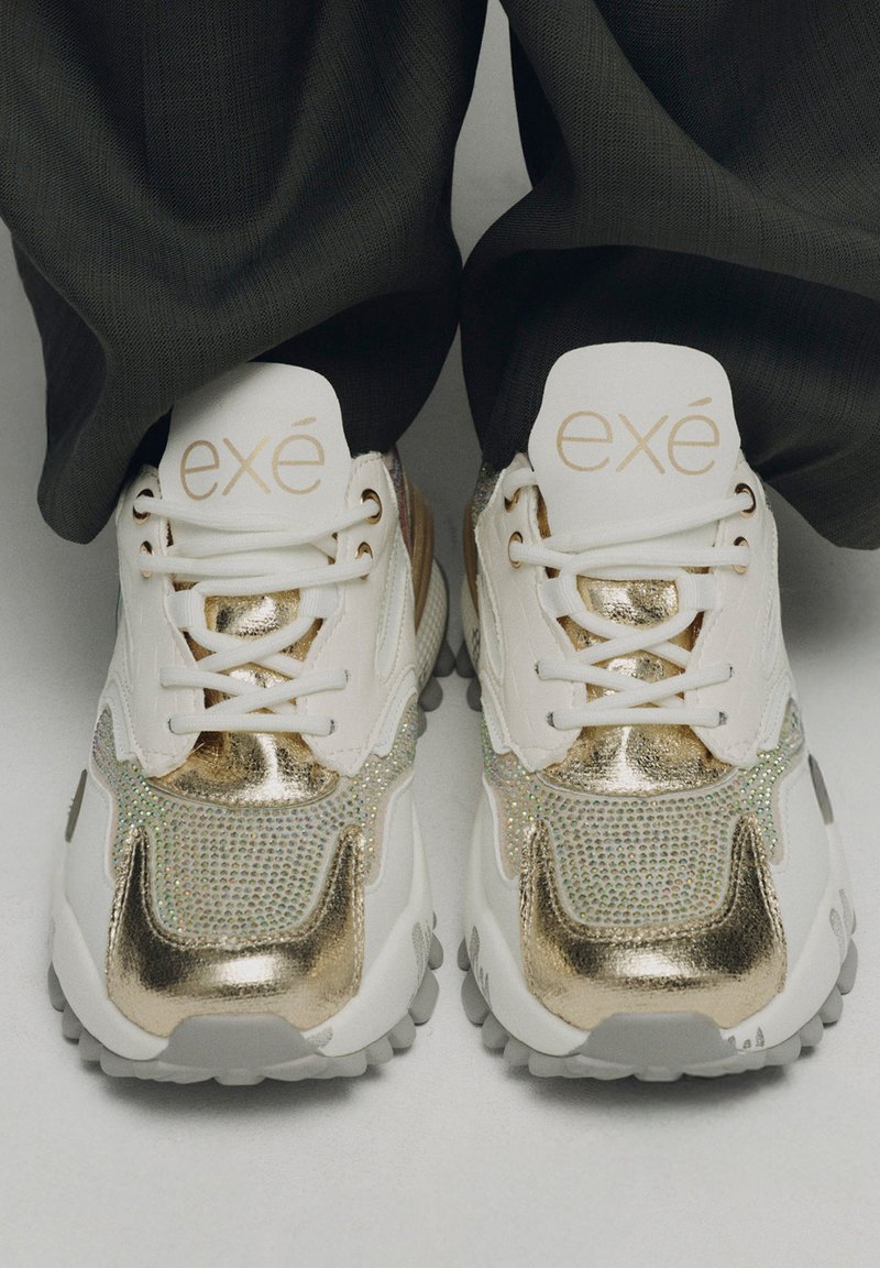 Zapatillas blancas con acentos de metal dorado y detalles iridiscentes, que cuentan con una suela acolchada y un diseño de cordones. Logotipo en la lengüeta.