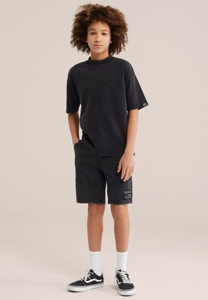 Ragazzo con capelli ricci in piedi, indossa una T-shirt nera oversize, pantaloncini neri con toppa, calzini bianchi e sneakers Vans nere.