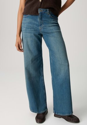 Frau trägt blaue Jeans mit weitem Bein, dunkelbraunes ärmelloses Oberteil und dunkelbraune Mokassins, steht vor einem schlichten Hintergrund.