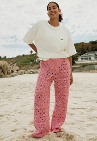 Felpa bianca oversize con la scritta "self" stampata in nero; pantaloni all'uncinetto rosa con motivi circolari, indossati su una spiaggia sabbiosa.