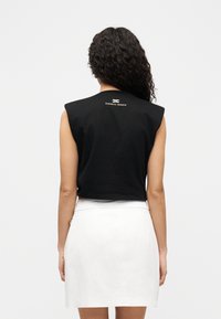 Μαύρο, κομμένο αμάνικο τοπ με δομημένους ώμους, που διαθέτει λεπτομέρεια λογότυπου στο πίσω μέρος. Συνδυάζεται με στενή λευκή φούστα.