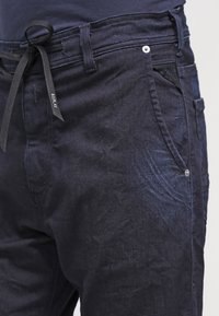 Pantaloni in denim blu navy con una texture liscia, dotati di cintura annodata, una tasca frontale e un dettaglio con bottone metallico in vita.