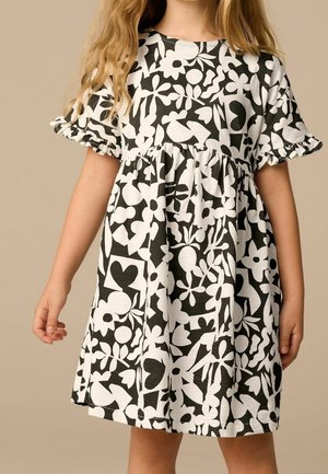 Enfant portant une robe noire et blanche à motifs floraux et abstraits avec des manches courtes volantées sur un fond beige uni.