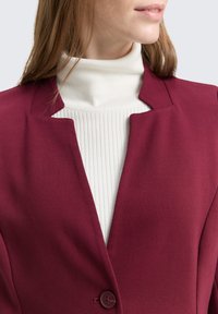 Bordo jakna od teksturiranog materijala sa strukturnim ovratnikom, s jednim dugmetom koje zatvara bijeli rebrasti džemper s visokim ovratnikom.