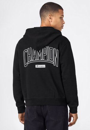 Jonge man in zijaanzicht, gekleed in een zwarte Champion-hoodie met grote witte tekst "CHAMPION" op de achterkant en donkere broek, voor een neutrale achtergrond.