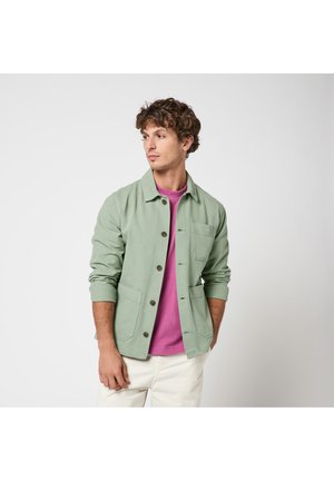 Joven con cabello rizado que lleva una chaqueta verde claro abotonada sobre una camisa morada y pantalones blancos, mirando hacia su izquierda sobre un fondo liso.