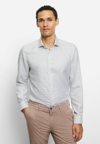 Hellgraues kariertes Hemd mit langen Ärmeln, Knopfleiste am Kragen und glattem Baumwollmischgewebe. Die Hose ist beige.