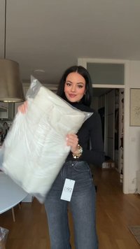 Fehér párnázott tárgy áttetsző műanyag zacskóban, amelyet egy fekete felsőt és sötét farmert viselő személy tart. Látni a "D92" felirattal ellátott címkét.