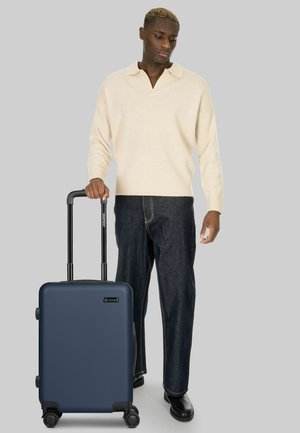 Homme en pull crème et jean foncé tenant la poignée d'une valise rigide bleu marine à quatre roues.