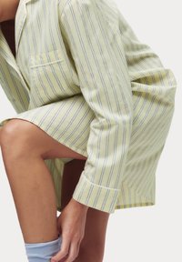 Conjunto de pijama a rayas amarillas que incluye una camisa con cuello, un bolsillo y mangas largas, acompañado de pantalones cortos hasta la rodilla. Textura de algodón suave.