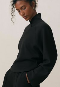 Czarny bluza z wysokim kołnierzem i długimi rękawami, o luźnym kroju i gładkiej fakturze. Subtelne przeszycia na ramionach.