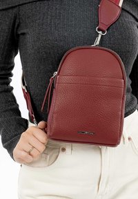 Červená kožená crossbody kabelka se zaobleným tvarem, texturovaným povrchem, zipovým uzávěrem a přední kapsou. Nastavitelný popruh s kovovými detaily.
