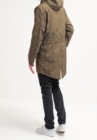Olivově zelený dlouhý kabát s kapucí, stažený v pase a se šikmými kapsami. Kombinován s černými džínami a bílými teniskami. Hladká, voděodolná látka.