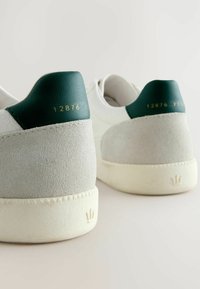 Baskets blanches en cuir avec des accents en suede vert au talon. Détails dorés embossés sur le talon. Tissu texturé et semelle blanche épaisse.