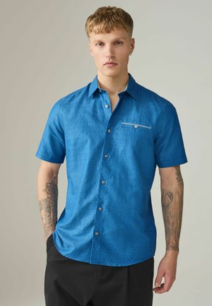 Ung mand med kort blondt hår iført en kortærmet blå mønstret skjorte med knapper og sorte bukser, stående foran en ensfarvet baggrund.